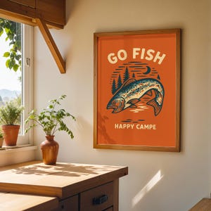 May include: A framed poster featuring a salmon illustration on an orange background. The poster has the text "GO FISH" above the fish and "HAPPY CAMPE" below. The fish is blue and white with a detailed design.