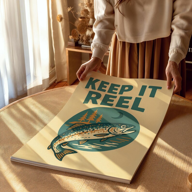 May include: A rectangular poster with the text "KEEP IT REEL" in teal. The poster features a teal circle with a fish illustration and trees. The background is a light beige color.
