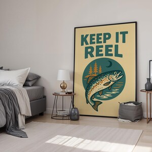 May include: A framed poster with the words "KEEP IT REEL" in teal lettering. The poster features a teal and brown fish illustration inside a teal circle with trees and a crescent moon. The background is a light yellow color.