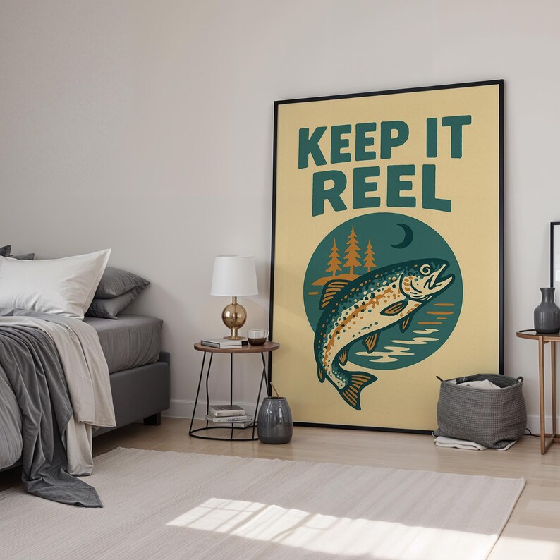 May include: A framed poster with the words "KEEP IT REEL" in teal lettering. The poster features a teal and brown fish illustration inside a teal circle with trees and a crescent moon. The background is a light yellow color.