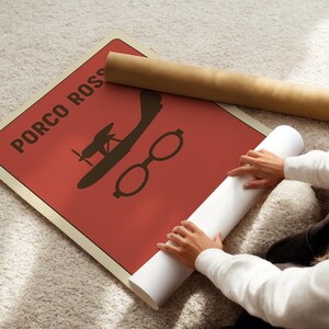 Puede incluir: Un p&oacute;ster rojo con el texto "PORCO ROSSO" y una silueta de un avi&oacute;n y gafas. Un p&oacute;ster enrollado y un papel blanco enrollado est&aacute;n junto al p&oacute;ster. El p&oacute;ster est&aacute; sobre una alfombra clara.