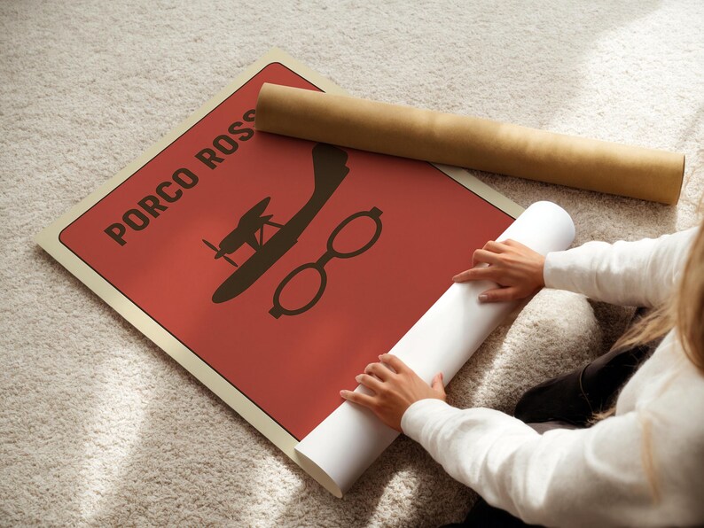 Puede incluir: Un p&oacute;ster rojo con el texto "PORCO ROSSO" y una silueta de un avi&oacute;n y gafas. Un p&oacute;ster enrollado y un papel blanco enrollado est&aacute;n junto al p&oacute;ster. El p&oacute;ster est&aacute; sobre una alfombra clara.