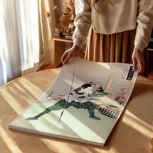 Könnte beinhalten: Ein Druck eines japanischen Holzschnittkunstwerks mit einem Krieger in grüner und schwarzer Kleidung, der ein Schwert und einen Speer schwingt. Das Kunstwerk hat einen hellen Hintergrund mit japanischem Text und wird von einer Person gehalten.
