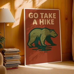 May include: A framed poster with the text "GO TAKE A HIKE" in cream-coloured letters above a green and brown bear illustration. The poster has a red background and a white frame, suitable for home decor.