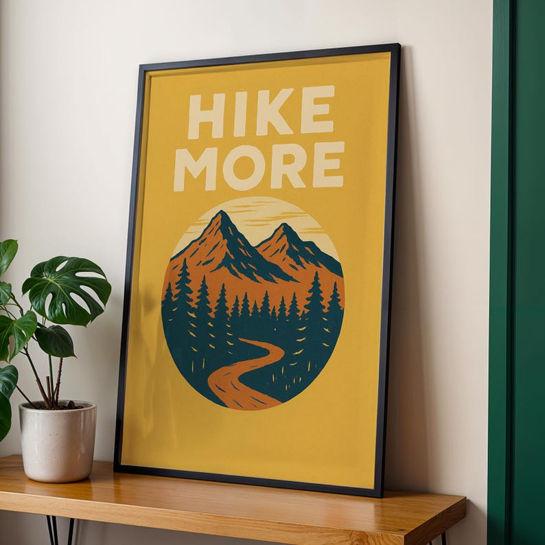 May include: Framed poster with the words "HIKE MORE" in white block letters. The poster features a mountain scene with orange and blue mountains, a forest, and a winding road, all set against a yellow background.