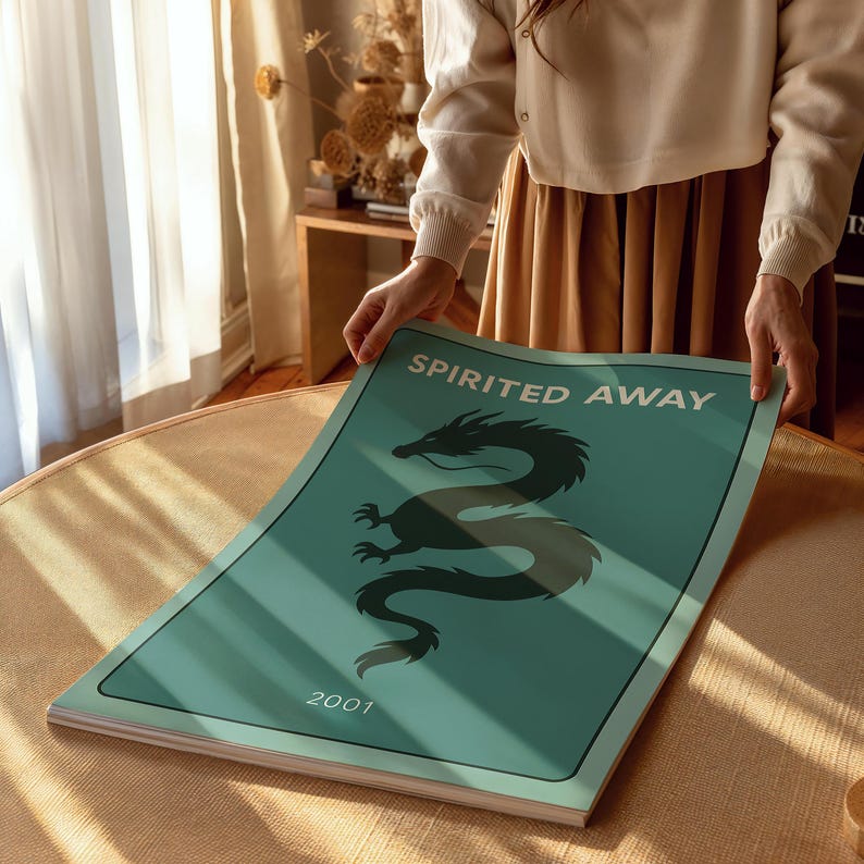 Puede incluir: Un p&oacute;ster turquesa con las palabras "Spirited Away" y el a&ntilde;o "2001" impresos. Una ilustraci&oacute;n de un drag&oacute;n negro est&aacute; centrada en el p&oacute;ster. El p&oacute;ster est&aacute; siendo sostenido por una persona.