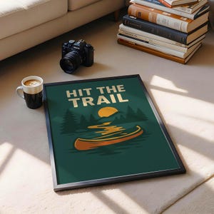 May include: Framed art print with the text "HIT THE TRAIL" above a canoe illustration. The artwork features a canoe on a lake with a sunset and trees, all set against a dark green background. A black camera and a coffee mug are nearby.