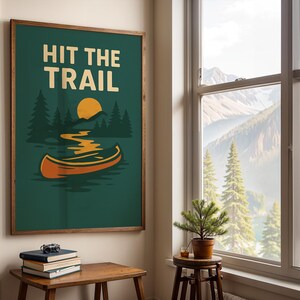 May include: A framed art print with the words "HIT THE TRAIL" in white block letters. The artwork features a canoe on a lake, a yellow sun, and green trees against a dark green background. The frame is brown.