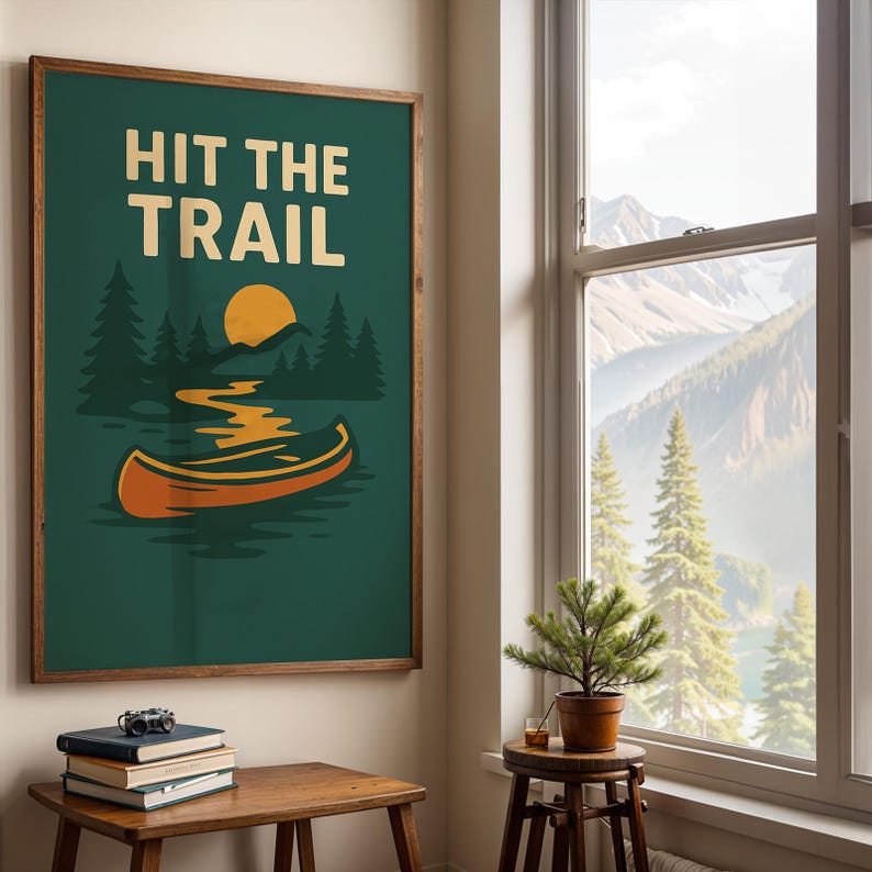 May include: A framed art print with the words "HIT THE TRAIL" in white block letters. The artwork features a canoe on a lake, a yellow sun, and green trees against a dark green background. The frame is brown.