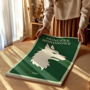 Puede incluir: Un p&oacute;ster verde con el texto "PRINCESS MONONOKE" y el a&ntilde;o "1997". El p&oacute;ster presenta una ilustraci&oacute;n de un lobo blanco. El p&oacute;ster est&aacute; siendo sostenido por una persona.