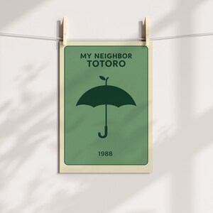 Puede incluir: Un p&oacute;ster verde con el texto "MY NEIGHBOR TOTORO" y el a&ntilde;o "1988". El p&oacute;ster presenta un gr&aacute;fico de paraguas verde oscuro. El p&oacute;ster est&aacute; sujeto por dos pinzas de madera en una cuerda blanca.