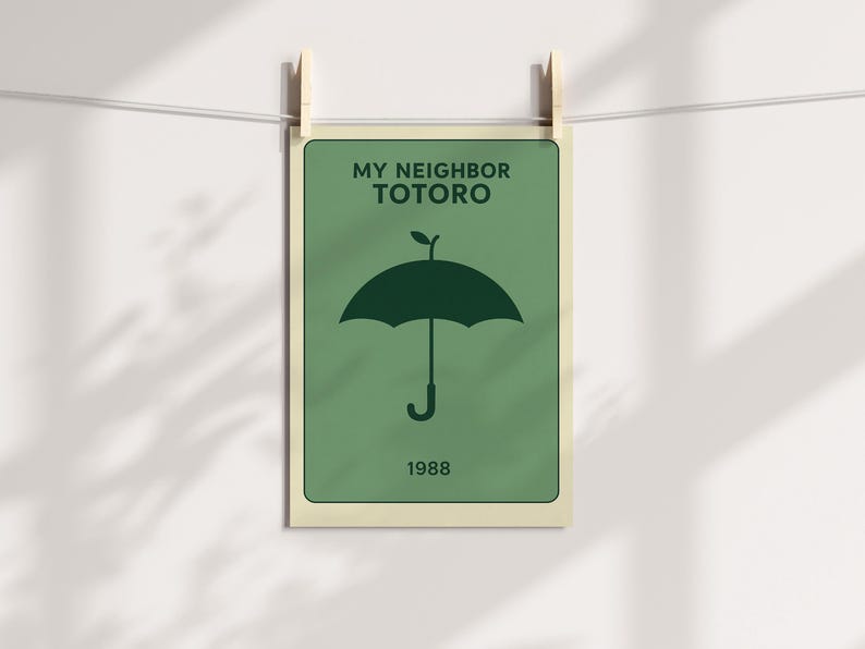 Puede incluir: Un p&oacute;ster verde con el texto "MY NEIGHBOR TOTORO" y el a&ntilde;o "1988". El p&oacute;ster presenta un gr&aacute;fico de paraguas verde oscuro. El p&oacute;ster est&aacute; sujeto por dos pinzas de madera en una cuerda blanca.