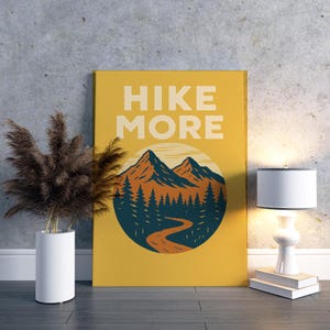 May include: A mustard yellow canvas print with the words "HIKE MORE" in white block letters. The print features a mountain scene with orange and blue mountains, a winding road, and pine trees. A white lamp and a vase with dried plants are next to the print.