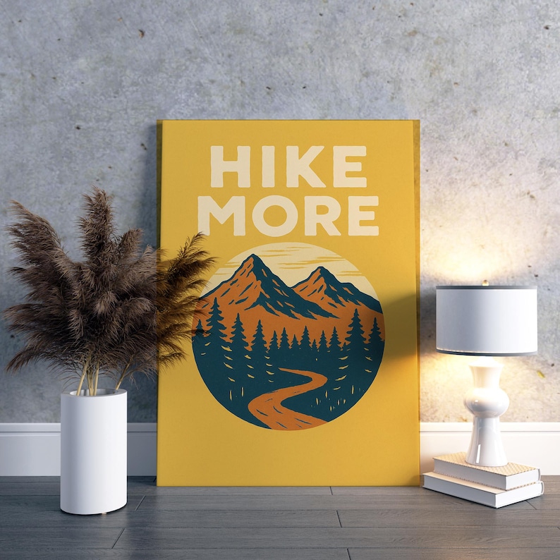 May include: A mustard yellow canvas print with the words "HIKE MORE" in white block letters. The print features a mountain scene with orange and blue mountains, a winding road, and pine trees. A white lamp and a vase with dried plants are next to the print.