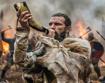 May include: A curved drinking horn featuring intricate gold-colored engravings of a bird, held by a person wearing a textured fur mantle over a light-colored tunic. The horn includes a thin leather strap and displays the text NORSE EMPIRE.