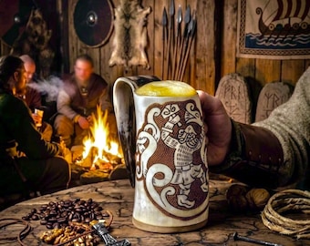 May include: A large, ornate beer mug filled with frothy beer, featuring a detailed Viking-style design in brown and cream. The mug is held in a person's hand, with a rustic wooden table and a cozy, dimly lit interior in the background.