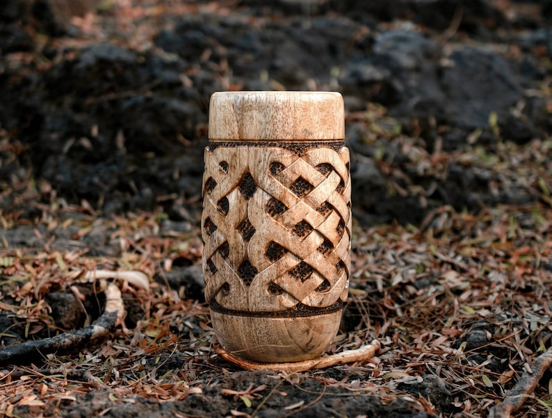 May include: A wooden cup with a carved Celtic knot design. The cup is light brown and has a smooth, polished finish.