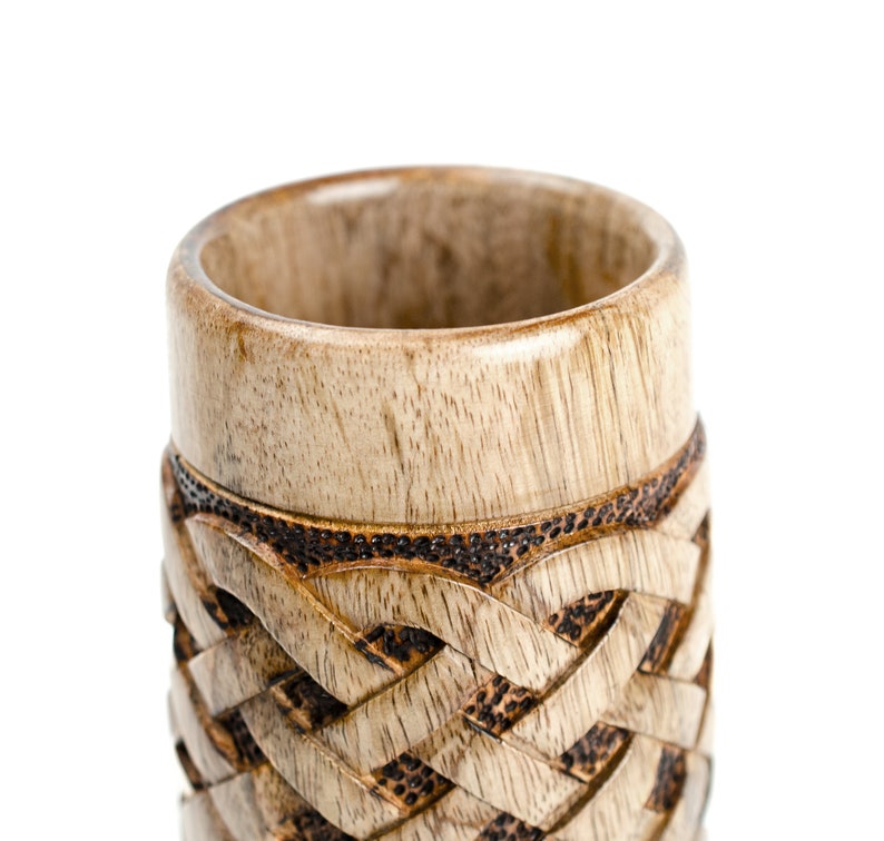 May include: A wooden cup with a carved Celtic knot design. The cup is made of light brown wood with dark brown accents.