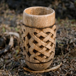 May include: A wooden cup with a carved Celtic knot design. The cup is light brown and has a smooth, polished finish.