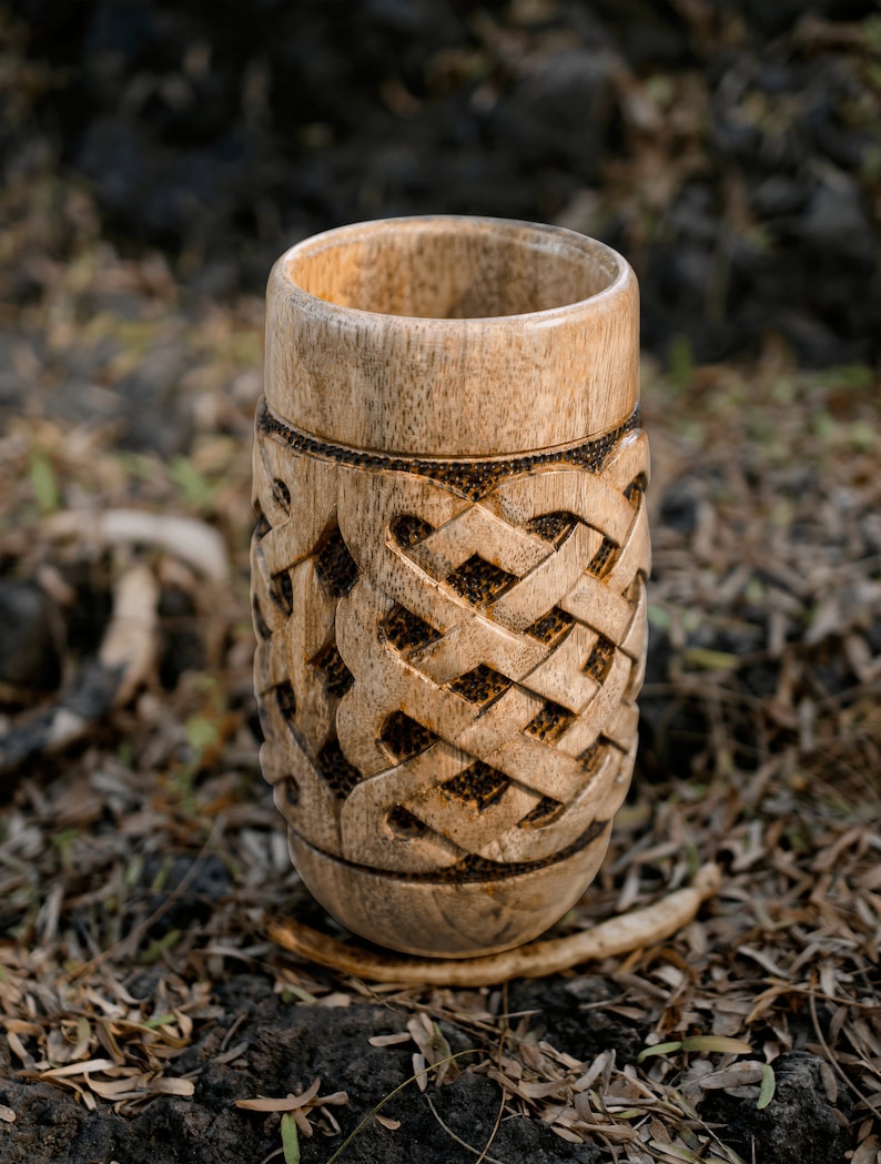 May include: A wooden cup with a carved Celtic knot design. The cup is light brown and has a smooth, polished finish.