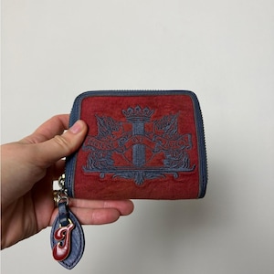 May include: Red zipped wallet with a blue trim and a matching blue and red charm. The wallet features a crest design with the words "HOUSE OF JUICY" in blue. The charm has a red "J" on a blue background.