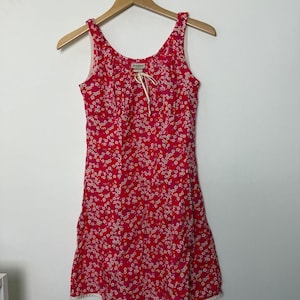 May include: A sleeveless dress featuring a vibrant red background with a dense pattern of small white and yellow flowers. The dress has a scoop neckline with a central white drawstring tie and a white hemline trim, hanging on a wooden hanger.