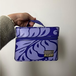 May include: A purple and lavender Roxy cosmetic bag with a floral design. The bag has a zipper closure, a handle, and the Roxy logo. The bag is held by a person.