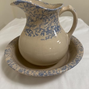 May include: A vintage cream-colored ceramic pitcher with a blue floral pattern and a matching saucer. The pitcher has a wide mouth and a handle.