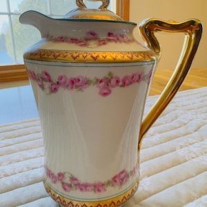 May include: A white porcelain pitcher with a gold handle and accents. The pitcher features a floral design of pink roses and gold trim. The lid has a gold handle. The pitcher is likely for serving beverages.