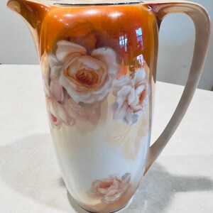 May include: A tall, vintage ceramic pitcher with a lid, handle, and spout. The pitcher features a cream-colored base with a floral design of pink and peach roses. The top is a gradient of orange and brown.