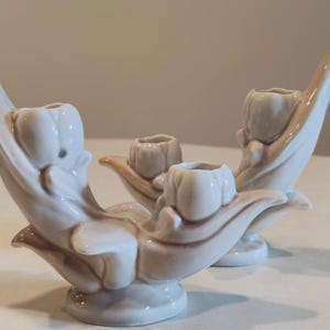 May include: Set of three ceramic candle holders. The candle holders are white with a beige accent. The design features a tulip-like shape with a curved, elongated stem. The candle holders are suitable for decorative purposes.