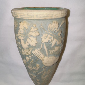 May include: A light blue ceramic wall pocket with a textured surface. The pocket features a relief design of leaves, vines, and a bird. The rim is a lighter color with a green interior. A decorative piece for home decor.