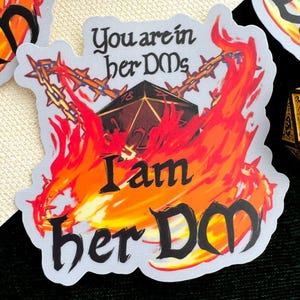 Puede incluir: Una pegatina con el texto "You are in her DMs, I am her DM" rodeada de llamas y un dado D20. La pegatina es blanca con gráficos negros y naranjas. Se ven varios dados dorados y negros.