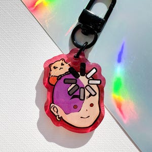 May include: A red acrylic keychain featuring a cartoon illustration of a person with purple hair and a small animal on top. The keychain has a black clasp and a small black ring. The background is a mix of white and iridescent colors.