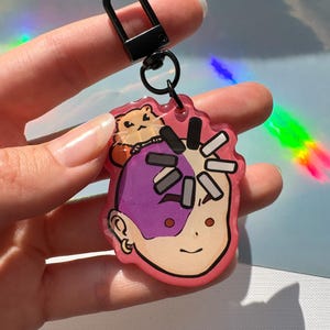 May include: A pink acrylic keychain featuring a cartoon character with purple hair and a hamster. The keychain has a black clasp and a small metal ring. The character has a smiling face and is outlined in black.