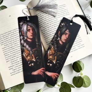 Peut inclure: Deux marque-pages noirs avec des illustrations de personnages fantastiques. Les personnages sont représentés avec des cheveux blancs, vêtus de vêtements sombres et tenant une épée. Les marque-pages sont décorés d'accents dorés.