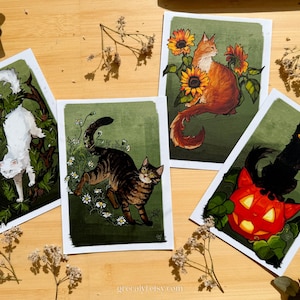 May include: Four art prints with cat illustrations. One shows a white cat on a branch, another a tabby cat among flowers, an orange cat with sunflowers, and a black cat on a jack-o'-lantern. The prints have a green background.
