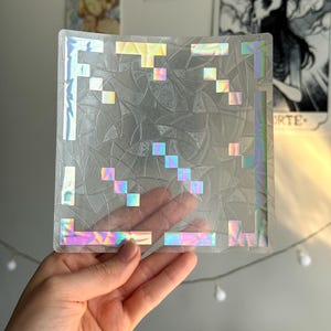 以下が含まれることがあります： ピクセル化されたデザインの透明な正方形のホログラフィックステッカー。ステッカーはテクスチャ加工された表面を持ち、虹色の色が反射するイリデセントアクセントがあります。ウェブサイトgrecaiyi.etsy.comが表示されています。