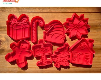 May include: A collection of red plastic Christmas-themed cookie cutters. The set includes shapes like a gift box, candy cane, elf hat, poinsettia, gingerbread man, North Pole sign, Christmas tree, and a house. The Bright Bakes Studio logo is in the top left corner.