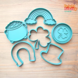 May include: A collection of turquoise cookie cutters in various shapes, including a rainbow, a pot of gold, a four-leaf clover, a horseshoe, and a leprechaun hat. The cookie cutters are arranged on a light wood surface. The Bright Bakes Studio logo is in the top right corner.