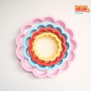 May include: A collection of four brightly coloured, scalloped-edge cookie cutters in pink, blue, red, and yellow, arranged in a nested circular pattern. The largest pink cutter is on the outside, with the smallest yellow cutter in the centre. The Bright Bakes Studio logo is visible.
