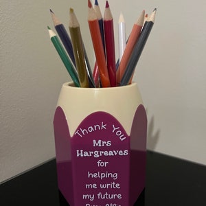 May include: A purple and cream pencil-shaped holder filled with coloured pencils. The holder has the text "Thank You Mrs Hargreaves for helping me write my future From Alfie". The pencils are various colours, including green, blue, red, and white.