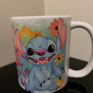 May include: A white ceramic mug with a colorful floral design featuring Stitch from Lilo & Stitch. Stitch is blue with big eyes and a toothy grin.