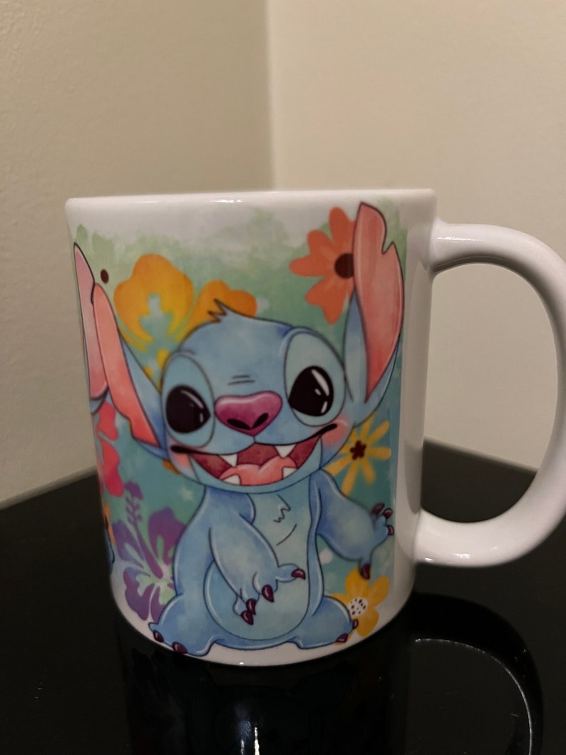 May include: A white ceramic mug with a colorful floral design featuring Stitch from Lilo & Stitch. Stitch is blue with big eyes and a toothy grin.