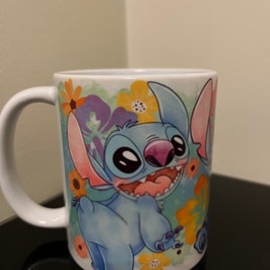 May include: A white ceramic mug with a blue and pink cartoon character of Stitch from Lilo & Stitch. The character is surrounded by colorful flowers and a watercolor background.