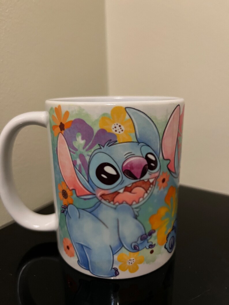 May include: A white ceramic mug with a blue and pink cartoon character of Stitch from Lilo & Stitch. The character is surrounded by colorful flowers and a watercolor background.