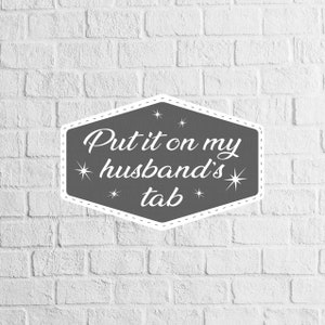 May include: A gray hexagonal sign with a white stitched border and white stars. The sign reads "Put it on my husband's tab".