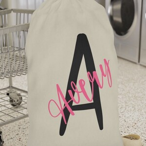May include: A pink canvas laundry bag with a black letter A and the name Avery written in pink script. The bag is sitting on a tiled floor in front of a washing machine.