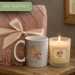 May include: A lit candle next to a personalized mug and a decorative blanket with a bow. The mug is gray with a gold letter "A" and floral design. The candle has a label with a floral design.