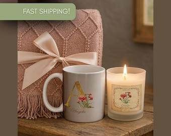 May include: A lit candle next to a personalized mug and a decorative blanket with a bow. The mug is gray with a gold letter "A" and floral design. The candle has a label with a floral design.
